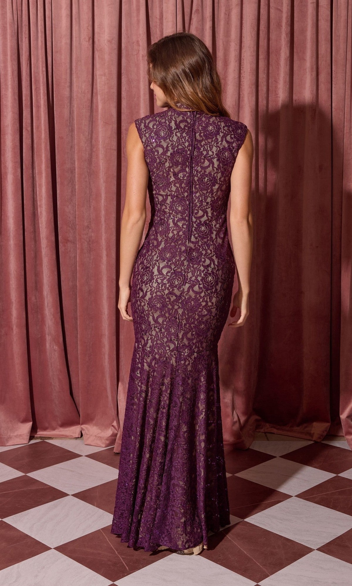 A woman faces away, wearing the Eureka 2061 Long Lace Formal Dress in purple, a sleeveless floor-length gown, standing before mauve curtains on a checkered floor.